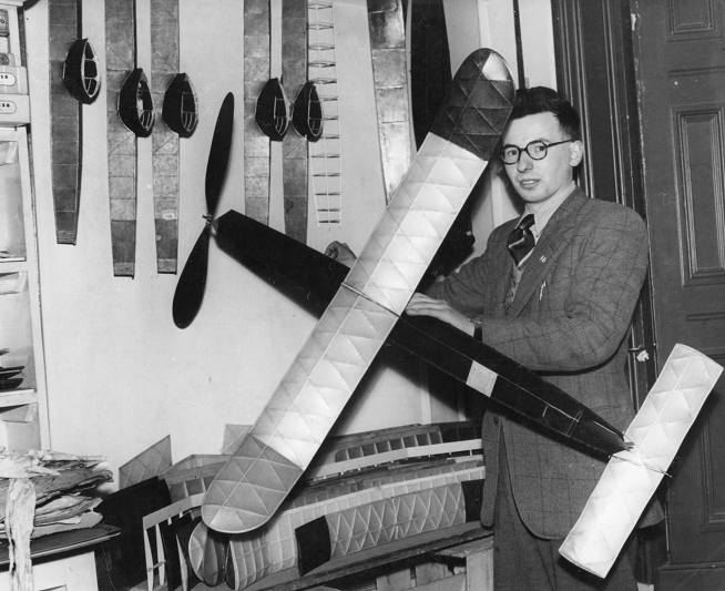 John in the 1940s, holding a large model, with various fuselages and wings arranged behind him.
